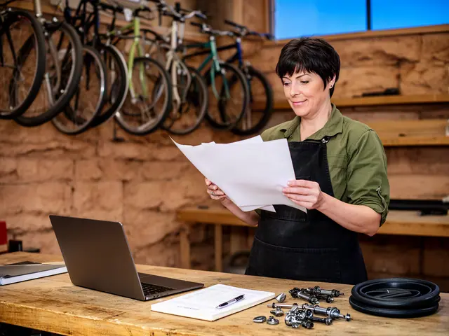 A woman in a green work shirt and black apron reads printed sheets beside a laptop in a bicycle shop.